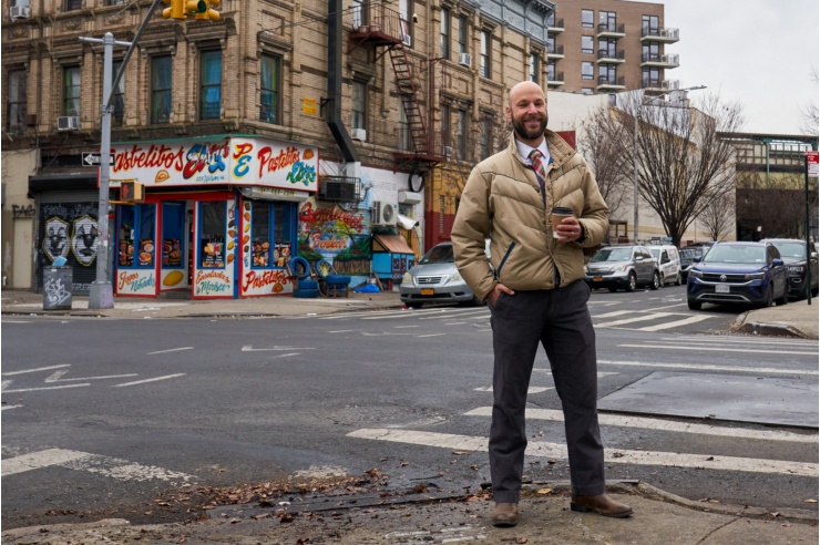 Ein Mann mit Glatze und Bart steht lächelnd an einer Straßenkreuzung in New York, im Hintergrund renovierungsbedürftige Altbauten.