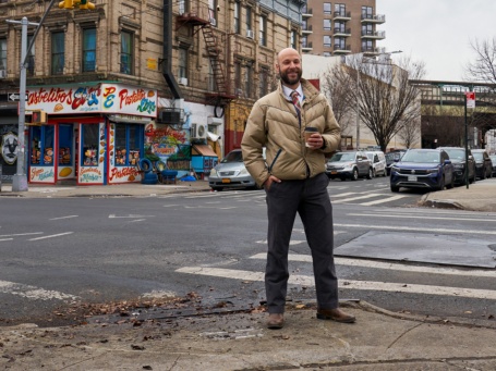Ein Mann mit Glatze und Bart steht lächelnd an einer Straßenkreuzung in New York, im Hintergrund renovierungsbedürftige Altbauten.
