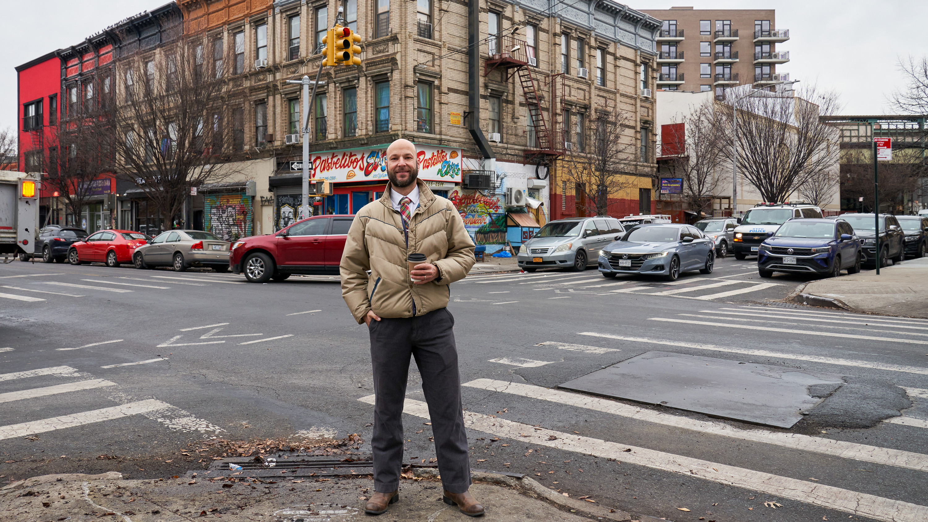 Ein Mann mit Glatze und Bart hält einen Kaffeebecher in der linken Hand, während er lächelnd am Straßenrand steht.