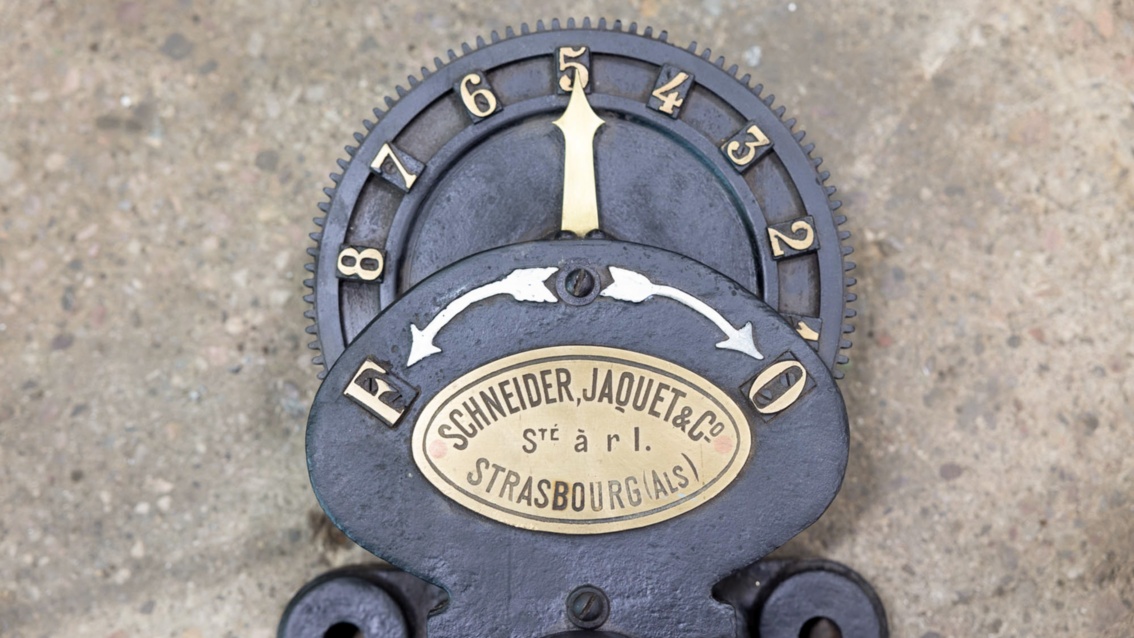 Eine alte schmiedeeiserne Anzeige an der Turbine: Pfeil, Zahlen und ein Emblem sind kunstvoll aus Messing gefertigt.