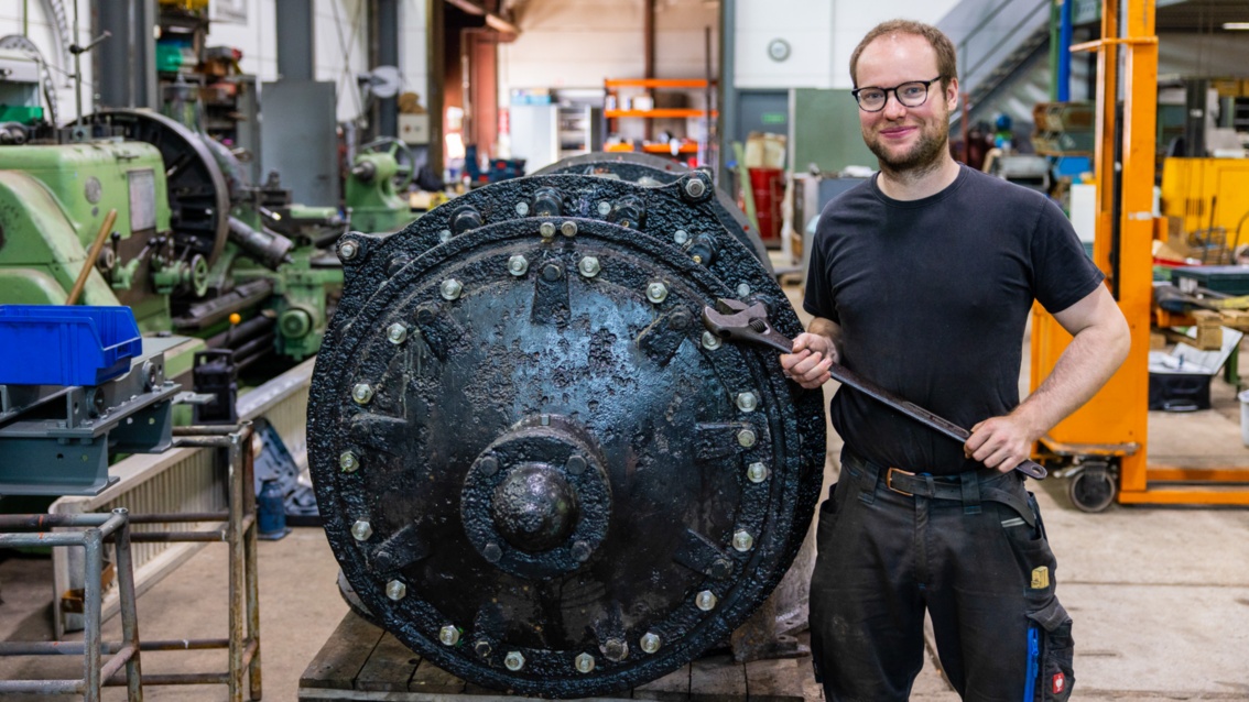 Ein Mann mit Brille steht stolz neben einer alten Maschine, in den Händen hält er einen großen Schraubenschlüssel. 