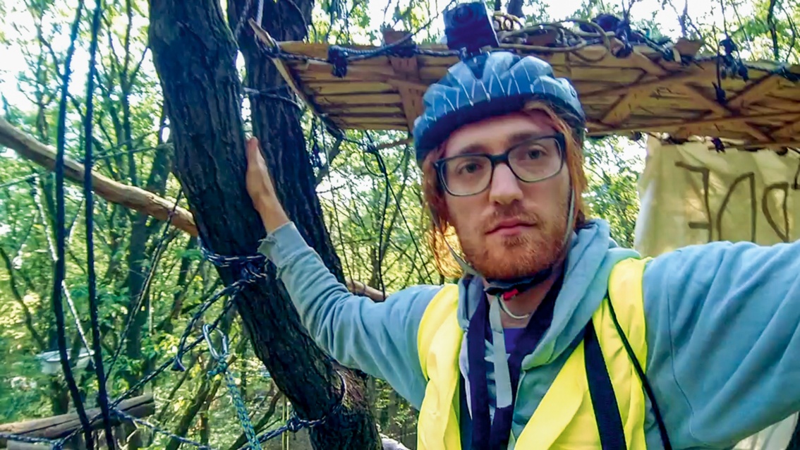 Ein junger Mann mit Dreitagebart, Brille und Helmkamera nimmt ein Selfie von sich in einem Baumhaus auf. 