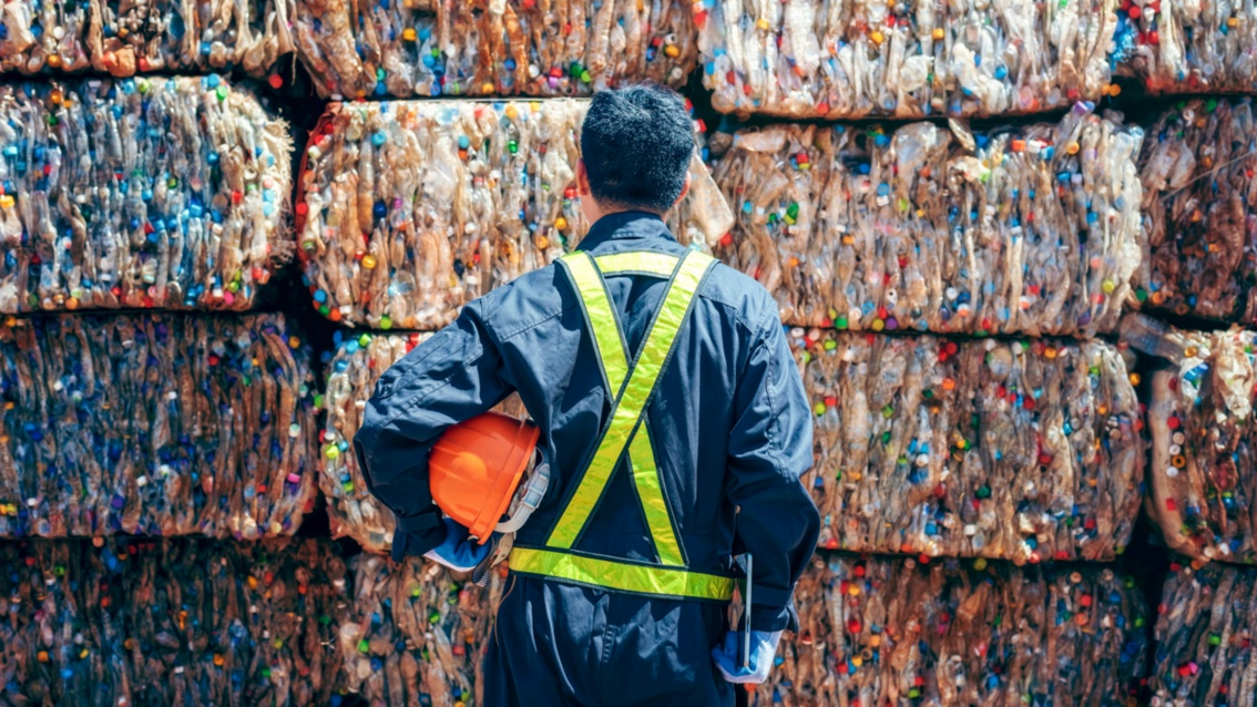 Rückansicht von einem Mann in Arbeitskleidung, der vor einer Wand aus großen Bündeln aus Kunststoffflaschen steht. 