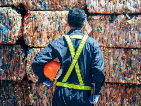 Rückansicht von einem Mann in Arbeitskleidung, der vor einer Wand aus großen Bündeln aus Kunststoffflaschen steht. 