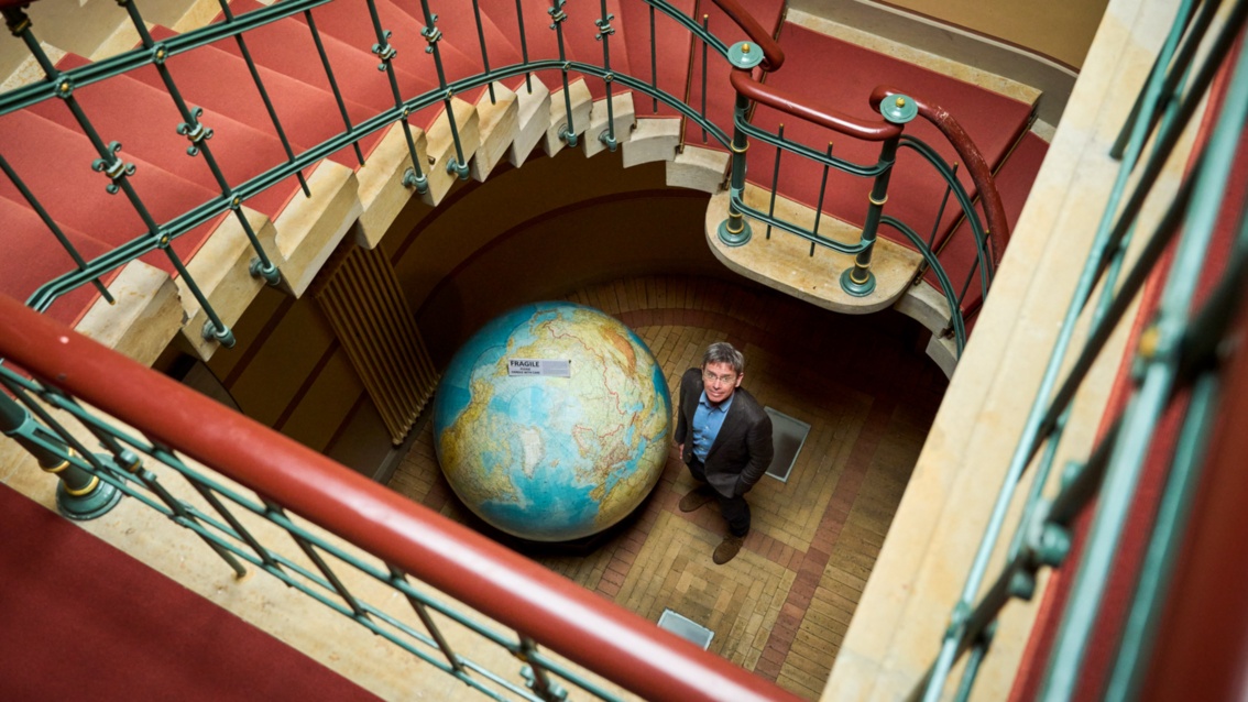 In einem großzügigen Treppenhaus steht ein Mann im Auge der Treppe, neben einem sehr großen Globus und schaut nach oben zur Fotografin.