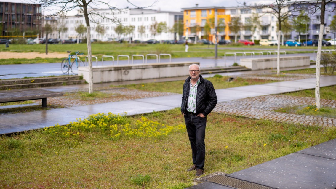 Carlo Becker, ein mittelalter Mann mit schütterem Haar, steht auf einer Grünfläche, im Hintergrund sind bunte Wohnblocks zu sehen.