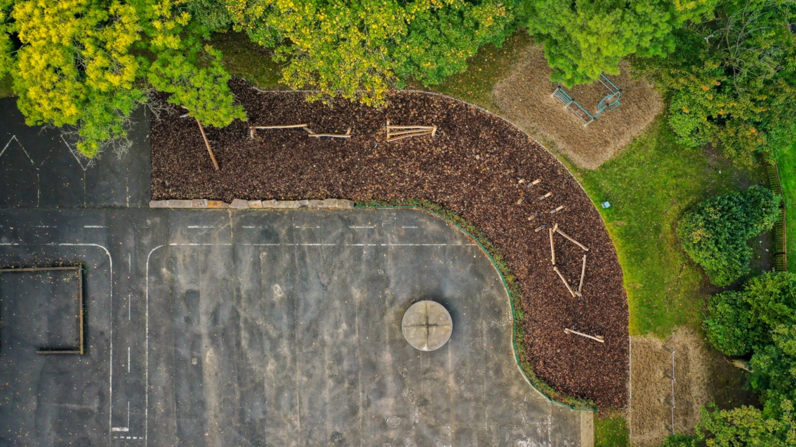 Drohnenaufnahme auf einen asphaltierten Platz, der im Halbrund gesäumt ist von Mulch und Grünpflanzen.