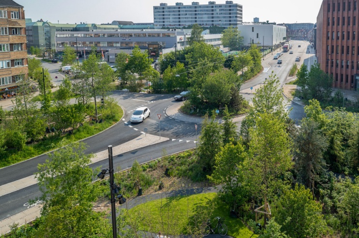 Ein Kreisverkehr und ein Straßenzug in einem städtischen Umfeld. Im Vordergrund bestimmen Bäume und Büsche das Bild.