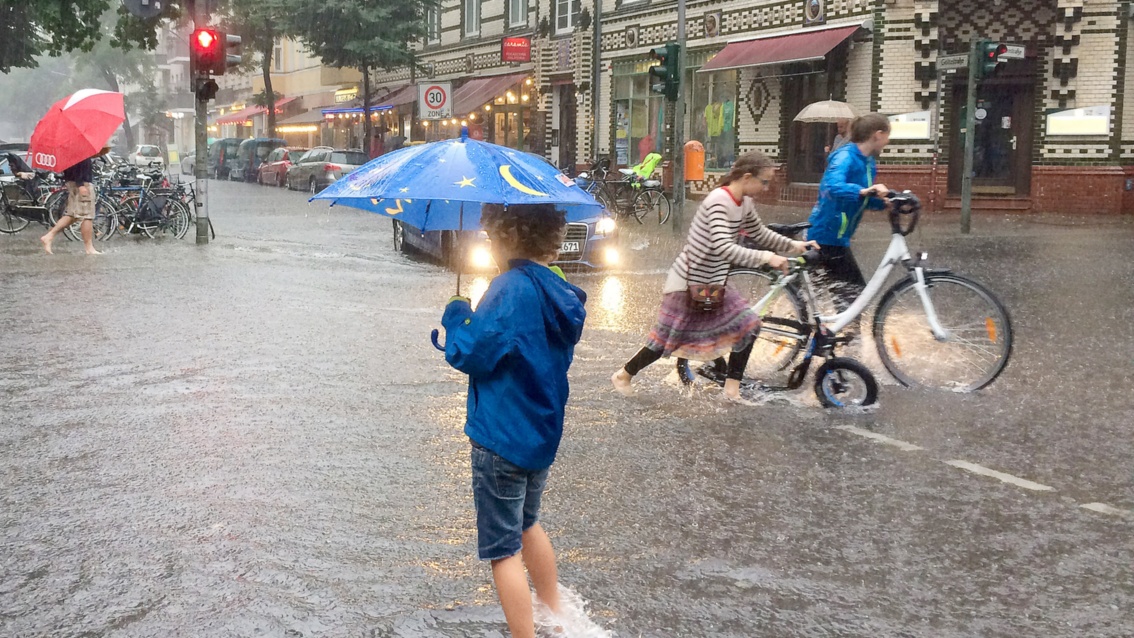 Eine städtische Straßenkreuzung ist knietief überflutet. Durch das Wasser watet ein Junge mit Regenschirm.