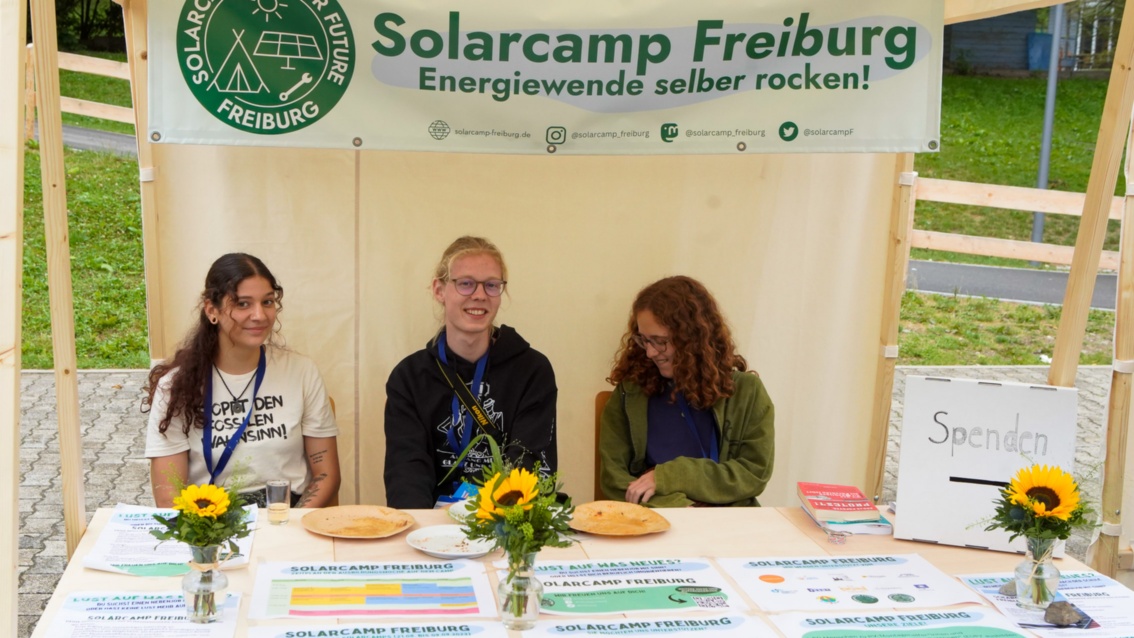 Ein junger Mann und zwei junge Frauen sitzen lächelnd hinter einem Stand mit der Aufschrift „Solarcamp-Freiburg“. 
