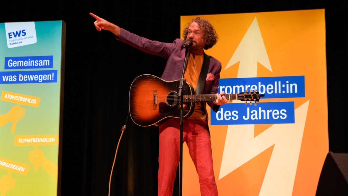 Ein Mann in lila Jackett und roter Hose: die linke Hand an der Gitarre, der rechte Zeigefinger in die Höhe gereckt.