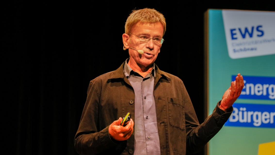 Ein Mann mit grau meliertem Haar und rahmenloser Brille hält einen Vortrag auf der Bühne.  