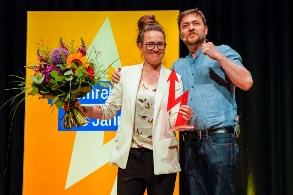 Katja Diehl steht mit einem großen Blumenstrauß und der Trophäe, einem roten Blitz zusammen mit Sebastian Sladek auf der Bühne.