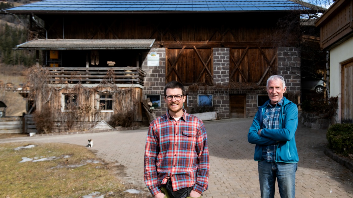 An elderly man stands in front of his homestead with his adult son, both wearing checked shirts. 