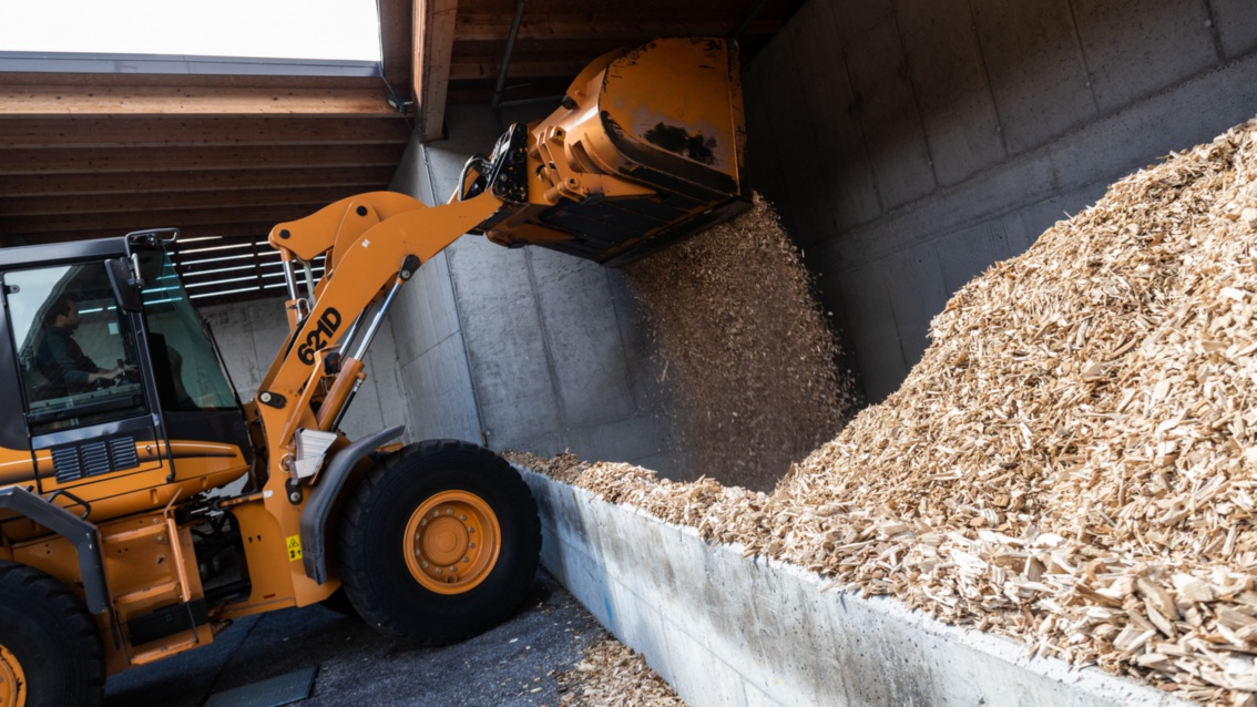 An orange excavator transports wood chips into a silo. 