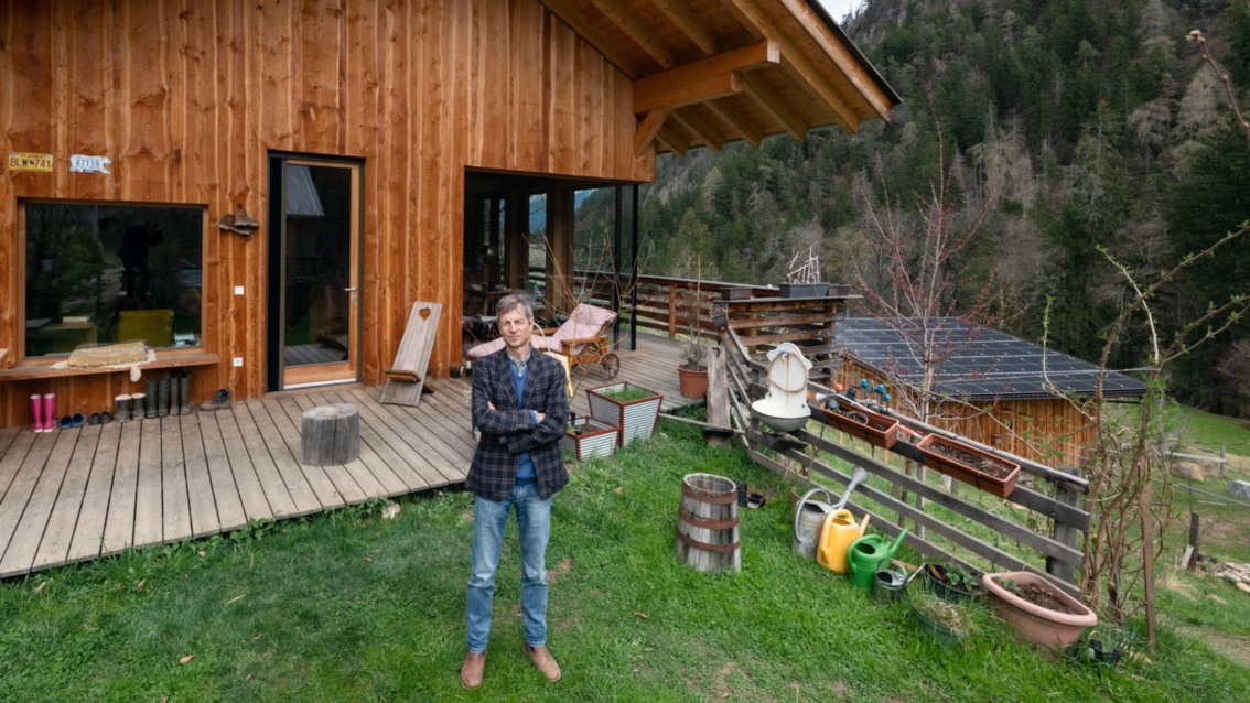 A man in a checked jacket stands in front of a modern wooden house and looks at the camera with his arms folded. 