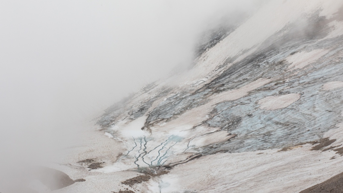 Vogelperspektive: Zwischen Schnee und Eis schlängeln sich kleine Bächlein den Hang herab, um in einer Nebelwand zu verschwinden.