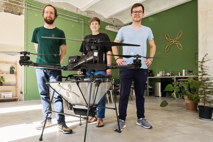 Drei junge Leute stehen  in einer Fabriketage stolz hinter einer etwa ein Meter hohen Drohne.
