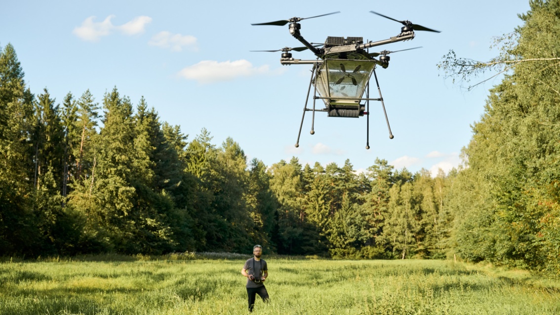 Eine Drohne schwebt über einer Waldlichtung – der Pilot steht wenige Meter entfernt.