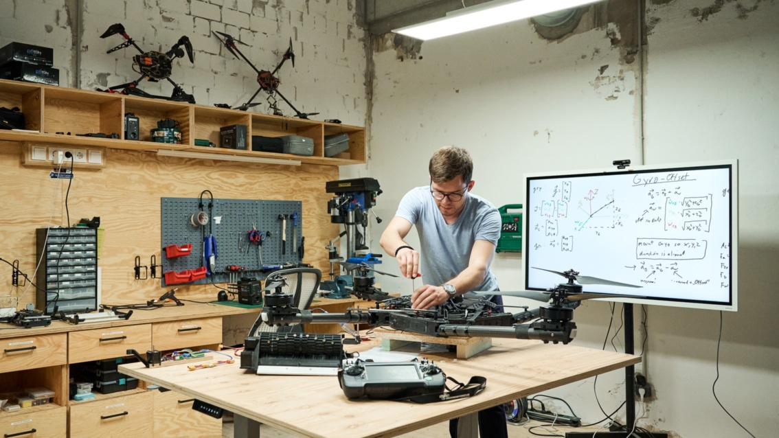 Ein Mann mit Brille arbeitet in einer Werkstatt an einer Drohne – auf einem Smartboard stehen Formeln und Skizzen.