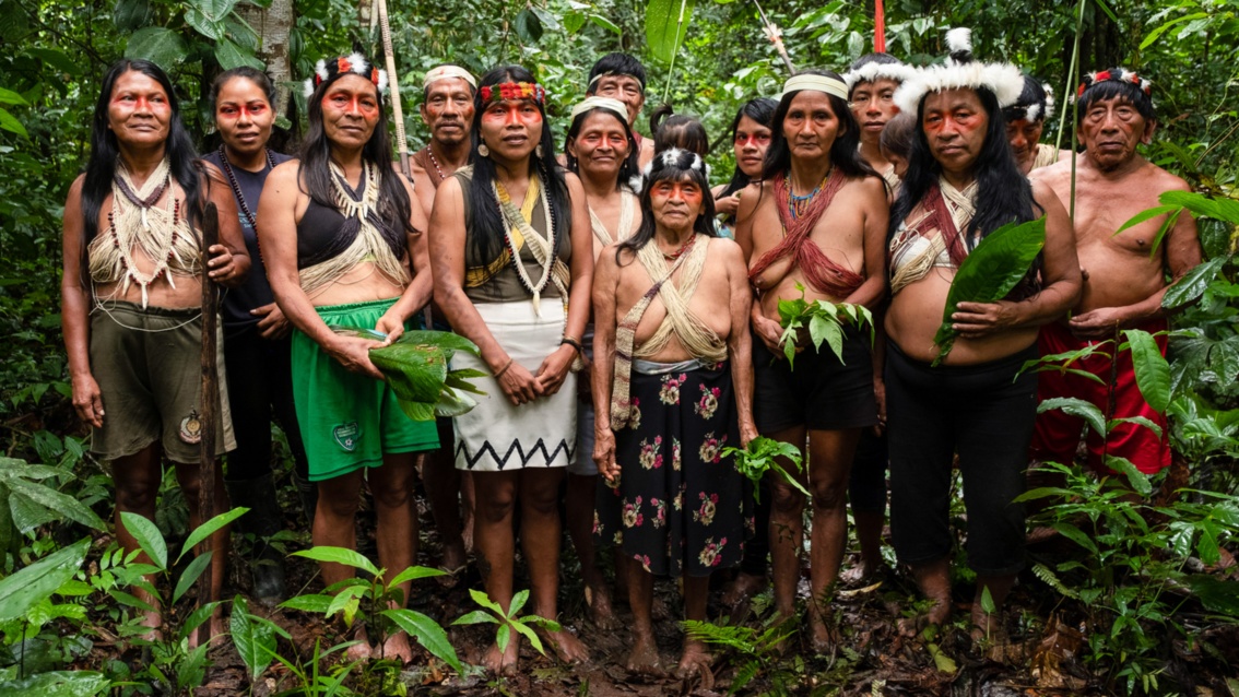 Eine Gruppe Indigener posiert in traditioneller Kleidung und mit Federschmuck – einige halten große Blätter.
