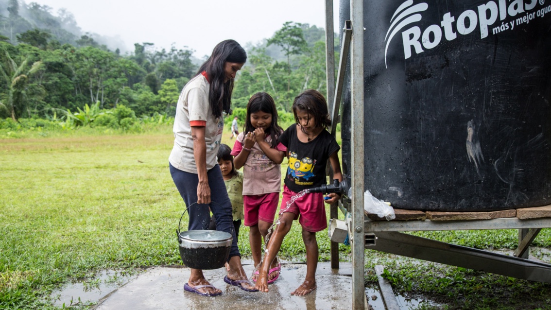 Eine Frau und drei Mädchen halten ihre Füße unter den Hahn eines Wassertanks, nachdem sie einen Topf befüllt haben.