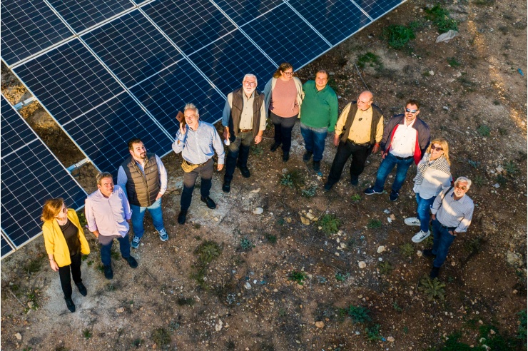 A drone photo of a group of thirteen people standing in a semicircle in front of a photovoltaic system.