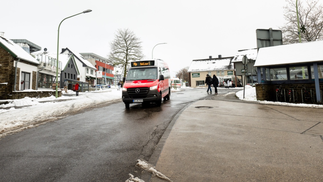 Ein kleiner rot-weißer Linienbus fährt durch eine verschneite Ortschaft mit Einfamilienhäusern. 