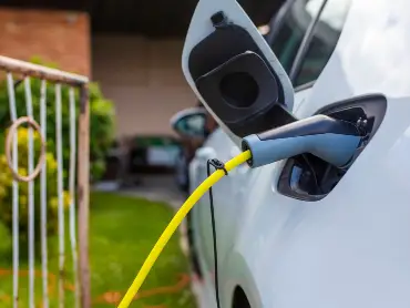 Ein Auto mit eingestecktem Ladekabel steht an einer Garageneinfahrt, im Hintergrund ist ein Wohnhaus mit Klinkerfassade zu erkennen.