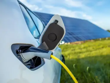 Vor einem Fld mit einer großen PV-Anlage steht ein weoßes Auto, das gerade mit Strom geladen wird.