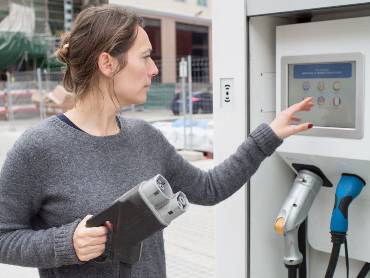 Eine junge Frau mit grauem Pullover steht an einer öffentlichen Ladesäule und tippt auf deren Display.