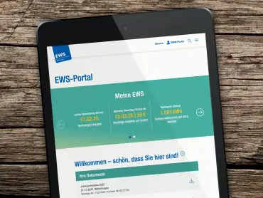 Auf einem groben Holztisch ein schwarzes Tablet, die aufgerufene Website ist die Start- und Willkommensseiteite des EWS-Portals