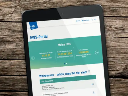 Auf einem groben Holztisch ein schwarzes Tablet, sie aufgerufene Website ist die Start- und Willkommensseiteite des EWS-Portals