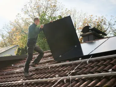 Ein Solateur auf einem Dach trägt ein PV-Modul, darauf sind Monatgeschienen, auf denen bereits viele Module installiert sind