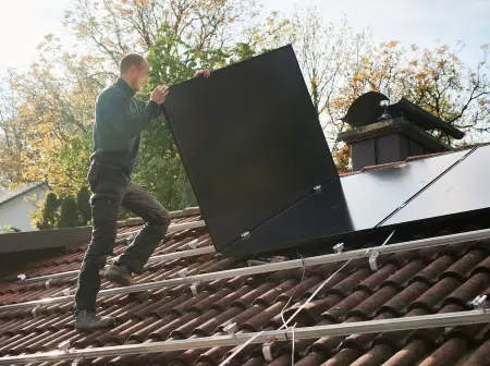 Ein Solateur auf einem Dach trägt ein PV-Modul, darauf sind Montageschienen, auf denen bereits viele Module installiert sind.