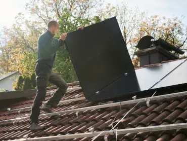 Ein Solateur auf einem Dach trägt ein PV-Modul, darauf sind Montageschienen, auf denen bereits viele Module installiert sind