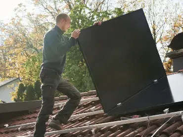 Ein Solateur auf einem Dach trägt ein PV-Modul, darauf sind Montageschienen, auf denen bereits viele Module installiert sind