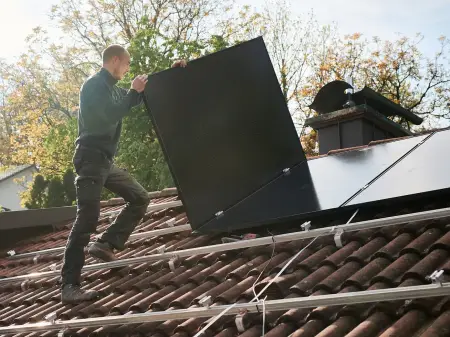 Ein Solateur auf einem Dach trägt ein PV-Modul, darauf sind Monatgeschienen, auf denen bereits viele Module installiert sind
