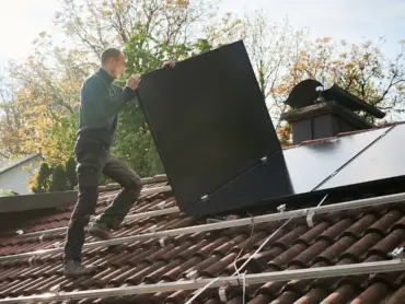 Ein Solateur auf einem Dach trägt ein PV-Modul, darauf sind Monatgeschienen, auf denen bereits viele Module installiert sind