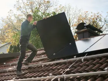 Ein Solateur auf einem Dach trägt ein PV-Modul, darauf sind Monatgeschienen, auf denen bereits viele Module installiert sind