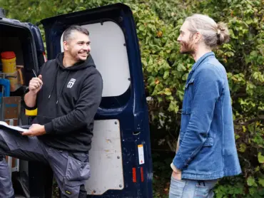 Vor der geöffneten Hecktüre seines Kleintramsporters steht ein Monteur, der gut gelaunt mit einem Mann spricht.