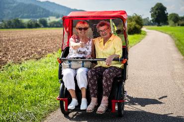 Zwei freundliche ältere Damen in einer Rikscha mit rotem Dach, eine hebt den Daumen