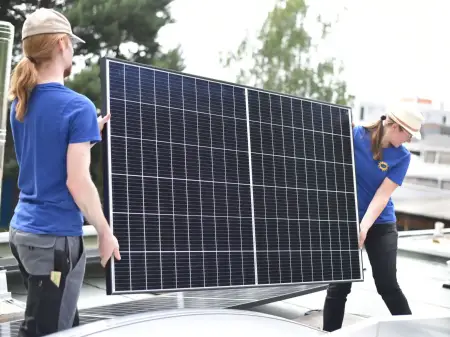 Zwei junge Menschen auf einem Hausdach tragen behutsam ein großes Photovoltaikmodul in Richtung eines im Hintergrund knienden Menschen.