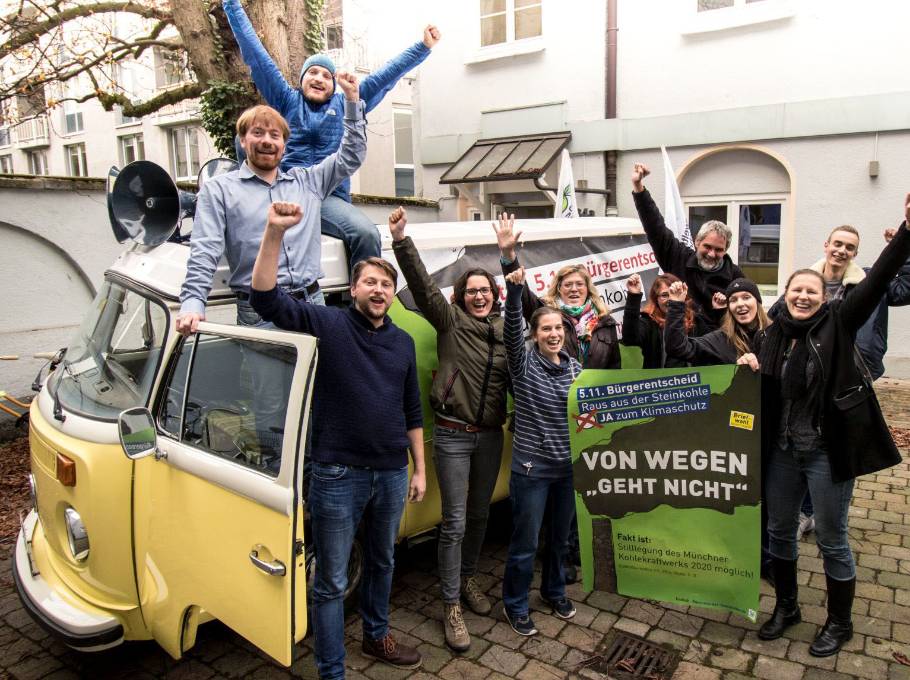 Vor einem Lautsprecherwagen reckt ein knappes Dutzend Bürger:innen siegessicher die Arme; die Plakaten, die sie halten, werben für einen Klimaentscheid.