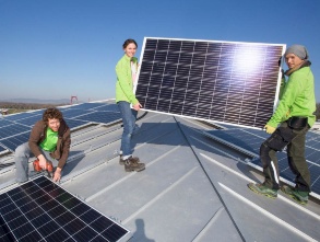 Auf dem Dach eines Mietshauses halten zwei Personen ein Solarmodul, eine dritte arbeitet an dem zugehörigen Montagegerüst.