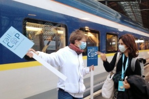 Vor einem Zug weist ein Mann mit einem «Rail to the COP»-Schild eine Reisende des Weges.