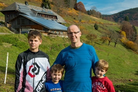 Ein Vater mit drei Söhnen steht vor einem Hang, im Hintergrund ist eine Scheune mit Solardach zu sehen