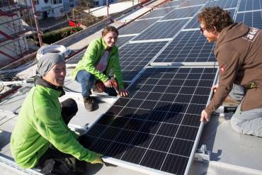 Drei Menschen installieren eine Solaranlage auf einem flachen Dach.