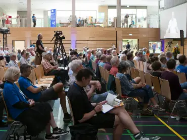 In einer Halle sitzen Teilnehmende an einer Veranstaltung, links im Bild eine Kamera auf einem Stativ samt Kameramann, der das Ereignis dokumentiert