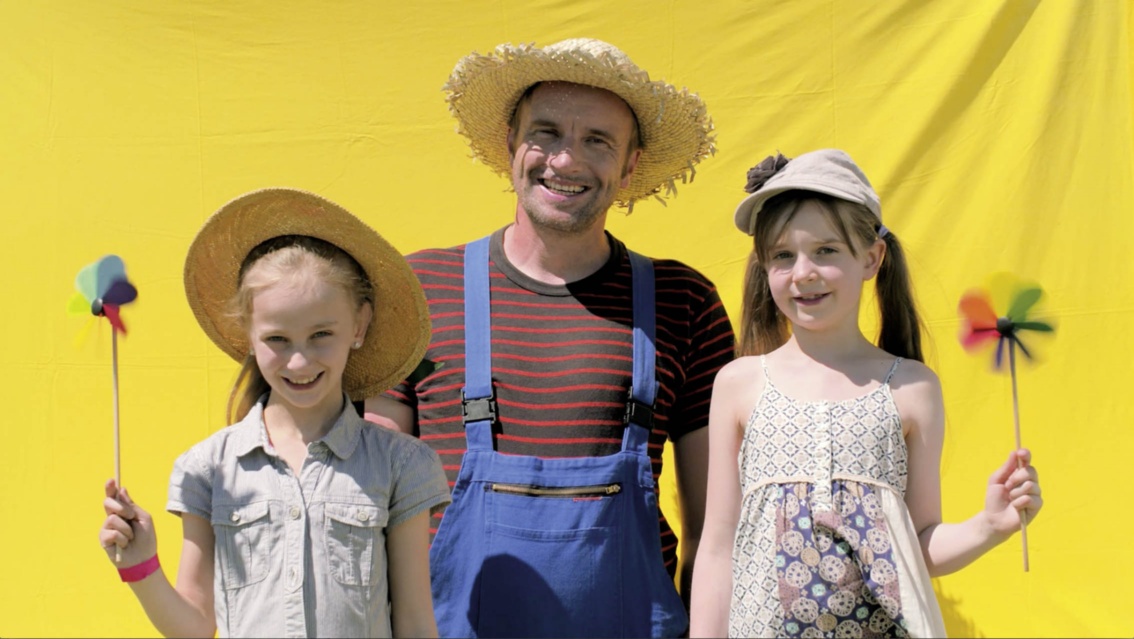 Ein lächelnder junger Mann mit Blaumann und zwei freundlich dreinblickende Mädchen mit Windrädchen in der Hand vor gelbem Hintergrund